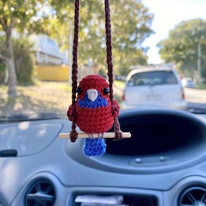 Pu&ograve; includere: Un piccolo ornamento a forma di uccellino all'uncinetto con un corpo rosso vivace, una macchia blu sul petto e una coda blu. L'uccellino ha occhi a perla neri e un becco bianco, ed &egrave; appollaiato su un bastoncino di legno orizzontale sospeso da due cordini intrecciati marroni.