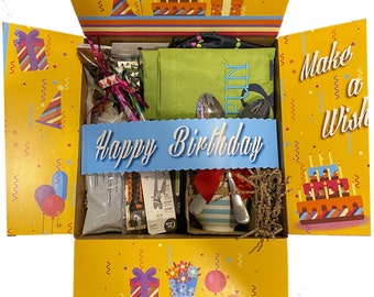 May include: A yellow and brown cardboard box with a birthday theme. The box is open and filled with various birthday party supplies, including a green apron, a blue and white striped plate, a red and white striped napkin, a silver spoon, and a silver fork. The box is decorated with colorful balloons, confetti, and the words "Happy Birthday" and "Make a Wish".