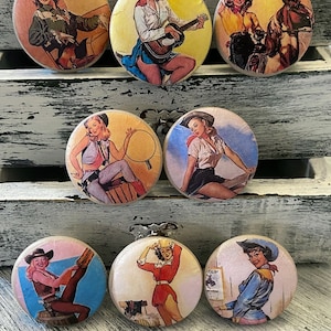 May include: Set of seven round wooden drawer pulls with colorful images of women in vintage western wear. The images include a woman playing a guitar, a woman riding a horse, a woman with a lasso, and a woman with a gun.