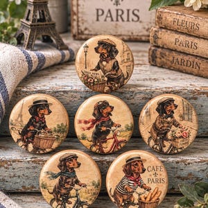 May include: A set of five round ceramic knobs featuring illustrations of dachshunds riding bicycles in various Parisian-themed outfits. Each knob displays a unique scene with a dog wearing hats, scarves, or vests, with some including baskets of flowers or bread.