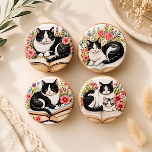 May include: Four round cookies with a cat theme. Each cookie features a black and white cat lying on an open book, surrounded by colorful flowers. The cookies have a light brown base and are decorated with intricate details.
