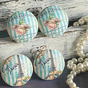 May include: Three round, white, wooden knobs with blue and white striped backgrounds. The knobs feature vintage-inspired designs with images of flowers, the Eiffel Tower, and the text "Cafes & Legumes" and "Cafe de Paris".
