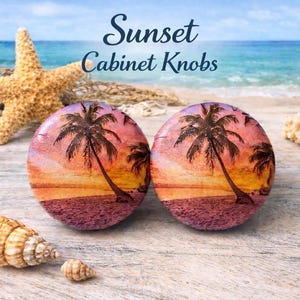 May include: Two round cabinet knobs with a sunset and palm tree design. The knobs feature a vibrant sunset with shades of orange, pink, and purple. The text "Sunset Cabinet Knobs" is displayed above the knobs.