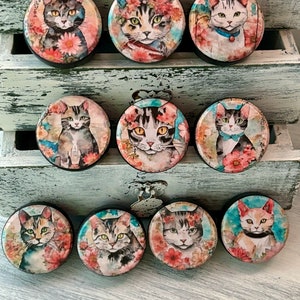 May include: Ten round, wooden knobs with colorful images of cats with floral accents. The knobs are painted in shades of blue, pink, green, and white.