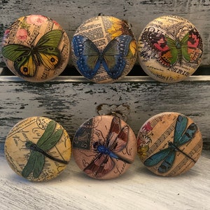 May include: Six round wooden knobs with vintage paper designs featuring butterflies and dragonflies. The knobs have a light brown background with a variety of colors and patterns.