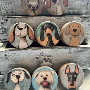 May include: A collection of nine round wooden cabinet knobs, each featuring a different dog breed illustration. The dogs are depicted in a whimsical, cartoon style with various colors and expressions. The knobs are set against a distressed white background.