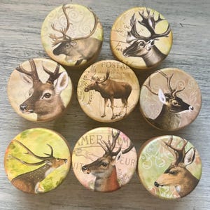 May include: A collection of eight round decorative cabinet knobs featuring detailed illustrations of deer and moose. The knobs have a vintage aesthetic with a muted color palette of beige, brown, and green, evoking a rustic, woodland theme.