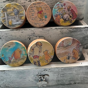 May include: Six round wooden knobs with Winnie the Pooh and friends designs. The knobs feature scenes from the Winnie the Pooh stories, including Winnie the Pooh, Piglet, Eeyore, and Tigger. The knobs are painted in a vintage style with muted colors and a distressed finish.