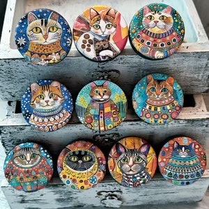 May include: Ten colorful round ceramic pinback buttons featuring different cats wearing sweaters and hats. The buttons are arranged in three rows on a white wooden surface.