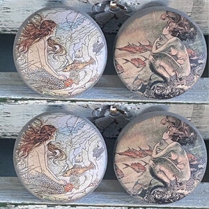 May include: Four round coasters with mermaid illustrations. Two coasters feature a mermaid with long brown hair, a fish, and aquatic plants. The other two show a mermaid with fish swimming around her. The coasters have a vintage, antique style.