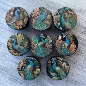May include: Eight round, black, decorative boxes with a peacock design. Each box features a peacock with blue and green feathers, gold accents, and a dark green and gold floral background.