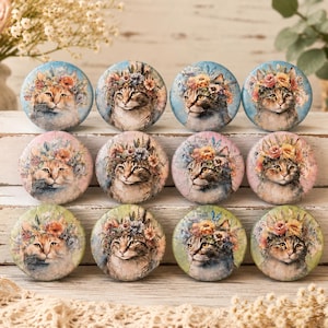 May include: A collection of twelve round cat-themed decorative magnets. Each magnet features a watercolor portrait of a cat wearing a floral crown, set against a pastel-colored background. The magnets are arranged on a white wooden surface.