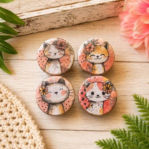May include: Four round cat-themed decorative items. Each features a smiling cat illustration with floral accents in shades of orange, pink, and white. The background has a dotted pattern. The items are arranged on a light-colored wooden surface.