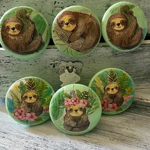 May include: Six round wooden knobs with painted images of sloths in a jungle setting. The sloths are brown with black eyes and are surrounded by green leaves and flowers.