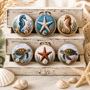 May include: Six round cookies with ocean-themed designs, including seahorses, starfish, and sea turtles. The cookies are decorated with blue, orange, and brown details, set on a white wooden display. Seashells and a starfish are in the foreground.