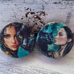 May include: Two round wooden coasters with a black cat and a woman's face with a cat's eye. The woman's face is on a blue and green background with a black cat's face partially covering her face. The other coaster has a black cat with a woman's face in the background. The woman's face is on a blue and green background.