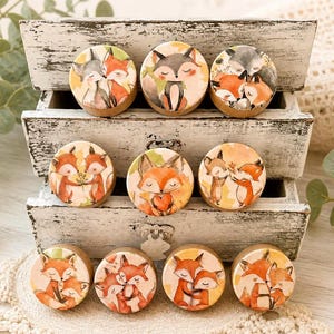 May include: A collection of round wooden decorative magnets featuring watercolor illustrations of foxes in various poses. The foxes are depicted in shades of orange, brown, gray, and white, set against a soft, warm background. The magnets are displayed in a rustic white wooden box.