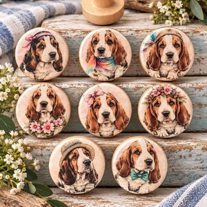 May include: A collection of eight round wooden cabinet knobs, each featuring a detailed illustration of a Basset Hound dog. The dogs are depicted with various accessories, including floral crowns, colorful scarves, bows, and hats, set against a soft background.