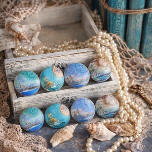 May include: A collection of nautical-themed round magnets with various designs, including ships and maps, displayed in a rustic wooden box. The magnets are in shades of blue, teal, and beige. The scene is decorated with seashells, pearls, and lace.