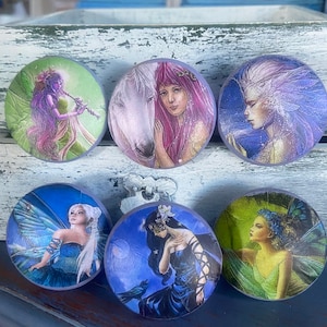 May include: Six round decorative magnets featuring fantasy art. The images depict fairies and a woman with a white horse. The color palette includes shades of purple, blue, green, and white. The magnets are arranged in two rows of three.