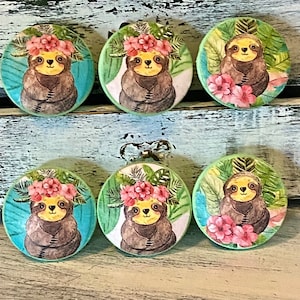 May include: Six round, green and blue, wooden drawer knobs with a sloth wearing a flower crown and surrounded by tropical leaves.