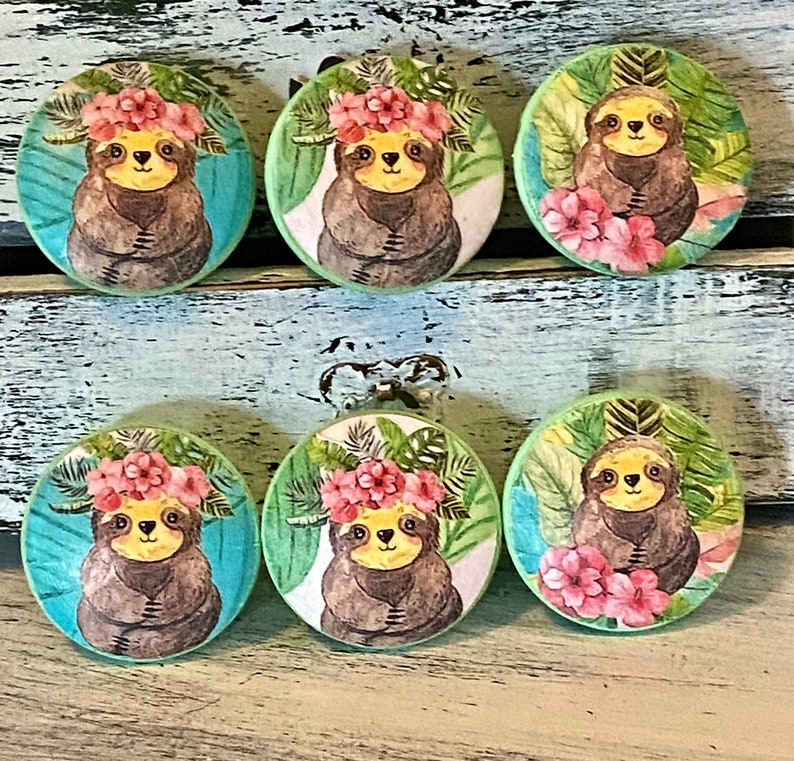 Op de afbeelding: Zes ronde, groene en blauwe, houten ladeknopjes met een luiaard die een bloemenkrans draagt en omringd is door tropische bladeren.