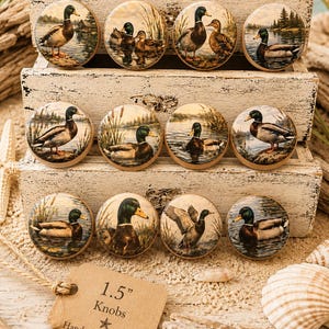 May include: A collection of 1.5-inch round knobs featuring detailed paintings of ducks in a natural lakeside setting. The knobs are displayed on a rustic wooden structure, with a tag that reads "Handmade with care."