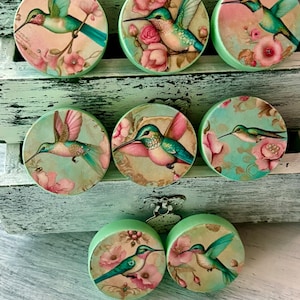 May include: Set of eight round wooden knobs with a green background and a floral design featuring hummingbirds. The hummingbirds are depicted in shades of blue and green with pink flowers.