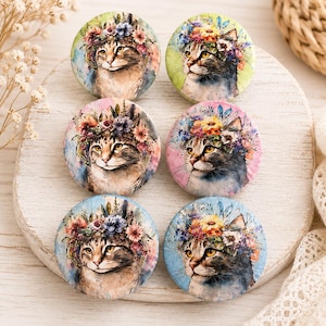 May include: Six circular buttons featuring a watercolor illustration of a cat wearing a colorful floral crown. Each button displays a cat with yellow eyes and a unique background color, including shades of green, pink, and blue, arranged in two columns.