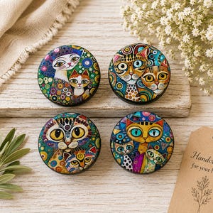 May include: Four circular cabinet knobs featuring colorful, intricate illustrations of cats with large eyes and patterned backgrounds. Each knob displays a unique artistic cat design with vibrant hues of blue, green, yellow, and orange in a mosaic style.