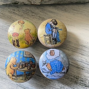 May include: Four round wooden knobs with Winnie the Pooh illustrations. The knobs feature scenes from the Winnie the Pooh stories, including Pooh Bear with honey pots, Pooh Bear sleeping, Piglet sitting on a chair, and Pooh Bear and Piglet walking in the woods.