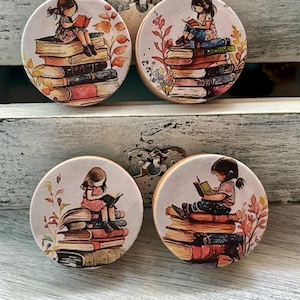 May include: Four round wooden knobs with a girl reading a book on a stack of books. The girl is sitting on the books and there are fall leaves and flowers around her. The knobs are painted in a white background with a light brown wood frame.