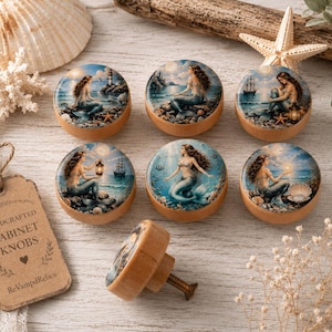 May include: A set of seven round wooden cabinet knobs, each featuring a unique circular illustration of a mermaid in a coastal setting. One knob is positioned upright to show the metal screw, while the others display detailed mermaid artwork.