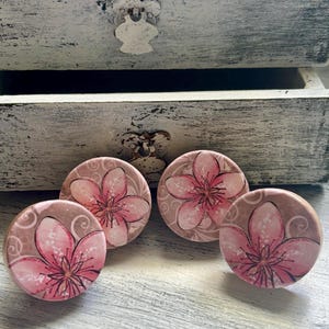 May include: Four round cabinet knobs with a floral design. Each knob features a pink flower with dark pink and black details on a brown background. The knobs are set against a distressed white and gray wooden drawer.