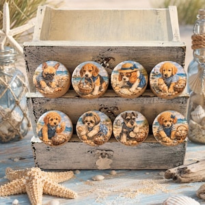 May include: Eight round wooden drawer knobs featuring colorful illustrations of various dog breeds on a beach. Each knob displays a different puppy wearing a blue striped shirt or accessory, with sand, seashells, and ocean water in the background.