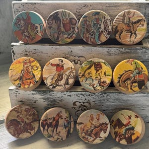 May include: A collection of eleven round wooden cabinet knobs featuring vintage cowboy and horse illustrations. The knobs display various scenes with cowboys, horses, and desert landscapes in a retro comic book style. The knobs are arranged in a white wooden box.