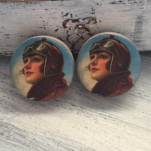 May include: Two round decorative cabinet knobs. Each knob features a vintage illustration of a woman wearing a leather aviator helmet and goggles, set against a blue sky background. The woman has red cheeks and lips.