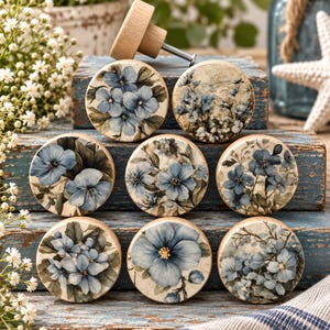 May include: A collection of eight round wooden cabinet knobs featuring various blue floral patterns on a cream background. Each knob has a flat circular face with detailed botanical illustrations of blue flowers and green leaves, and one knob shows the metal screw.
