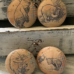 May include: Four round wooden cabinet knobs featuring Winnie the Pooh illustrations. Each knob has a different black and white drawing of Pooh Bear and friends. The knobs are attached to a white, distressed wooden surface.