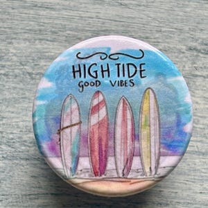 May include: A round, colorful button with a beach theme. The button features four surfboards in shades of pink, blue, and yellow on a sandy beach. The text "HIGH TIDE good VIBES" is written across the top in black script.