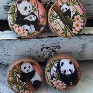May include: Four round wooden earrings with a pink background and images of pandas in a bamboo forest. The pandas are black and white and some have pink flowers around them.