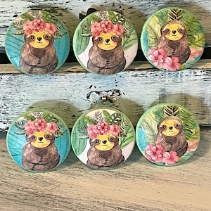 Op de afbeelding: Zes ronde houten knoppen met een tropisch luiaardontwerp. Elke knop heeft een luiaard die een roze bloemenkroon draagt en omringd is door groene bladeren.