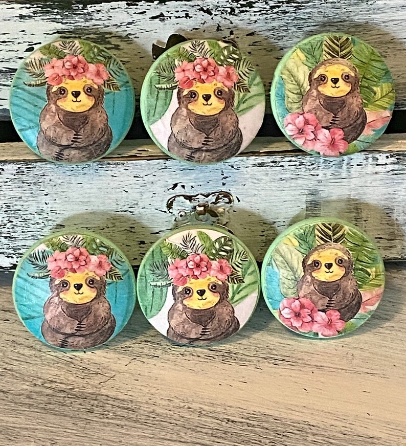 Op de afbeelding: Zes ronde houten knoppen met een tropisch luiaardontwerp. Elke knop heeft een luiaard die een roze bloemenkroon draagt en omringd is door groene bladeren.