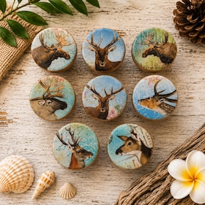 May include: A collection of nine round decorative cabinet knobs featuring detailed illustrations of deer and moose. Each knob has a unique design with a vintage aesthetic, set against a light blue, green, and floral background. The knobs are arranged on a light wood surface.
