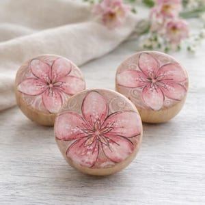 May include: Three wooden knobs with a floral design. Each knob features a pink flower with detailed petals and a light brown background. The knobs are arranged on a light-colored wooden surface, with a soft, neutral-colored cloth in the background.