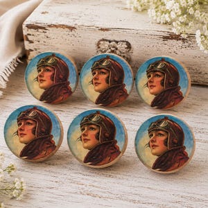 May include: A set of six round wooden drawer knobs, each featuring a printed illustration of a person wearing a vintage brown leather aviator helmet with goggles, set against a light blue sky background with a warm, golden-toned horizon.