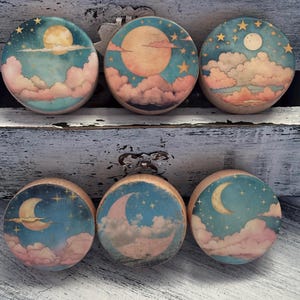 May include: Six round wooden decorative pieces with celestial designs. Each piece features a different moon phase, clouds, and stars in shades of blue, pink, and yellow. The pieces are arranged on a distressed white surface.