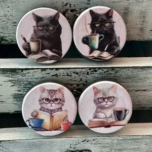 May include: Four round ceramic coasters featuring illustrations of cats wearing glasses and reading books. Each coaster has a different cat with a different book and a cup of coffee or tea.
