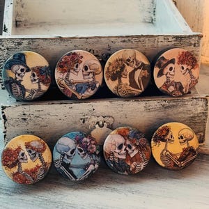 May include: Eight round wooden ornaments with colorful images of skeletons in various poses. The skeletons are wearing flowers and some have hats. The ornaments are arranged in two rows on a wooden surface.