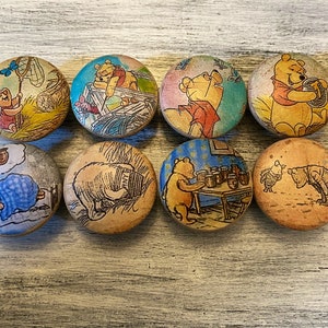 May include: Eight round wooden cabinet knobs featuring illustrations of Winnie the Pooh and friends. The knobs display various scenes with characters like Piglet, Pooh, and Eeyore, in a variety of colors including yellow, blue, and pink.